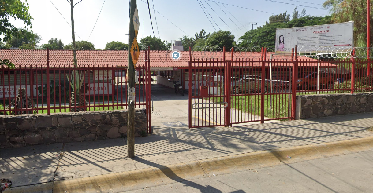 Fotografía en la que se muestra la entrada del plantel, conformada por una reja de color rojo. Detrás de la reja hay un pasillo que lleva a las instalaciones.
